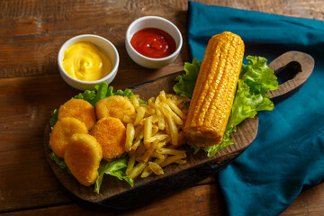 Nuggets, french fries and corn on the cob on a plank salad on a blue cloth.