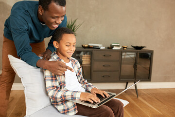 Father and son relationships. Picture of African American young smiling man standing behind boy of 7-8 years-old sitting in chair with laptop on knees, pointing at screen, helping him with homework