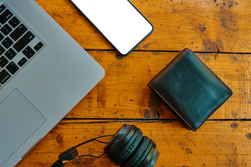 Top view of laptop, headphones, smartphone, and wallet on wooden table. Freelancer's work surface. Blank mobile phone screen to insert some text. Modern devices for business jobs and entertainment.