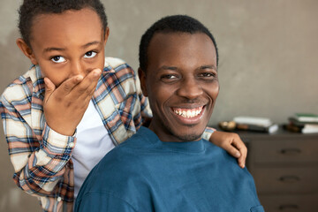 Close-up portrait of cute charismatic child covering mouth and nose holding hand over dad's shoulder, dark-skinned laughing father looking at camera, both in perfect mood isolated over room interior