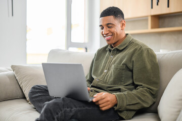 Optimistic man in casual wear using laptop while sitting on the couch. Young male student watching webinars, educational courses, learning on the distance, typing emails