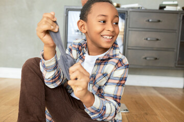 Charismatic good-looking cute boy in plaid shirt taking off or putting on grey sock, sitting on...