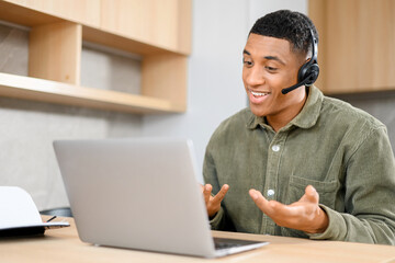 Concentrated young guy in smart casual wear is using headset and laptop for online communication, supporting, selling. Black guy sitting at the office desk