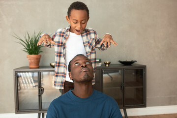 Father and son playing, dark-skinned dad sitting on floor, looking up at his boy's hands, who...