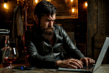 Obraz premium Bearded man drink cognac. Drunk attractive handsome sad pensive man in black jacket, business man drinking whiskey in front of laptop at loft work place.
