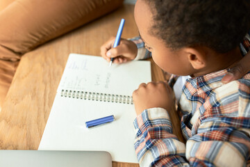 Upper view cropped shot of dark-skinned curly haired boy doing sums in his copy book, writing down...