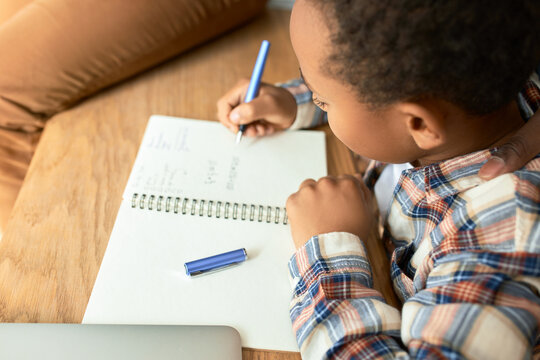 Upper view cropped shot of dark-skinned curly haired boy doing sums in his copy book, writing down letters and figures, sitting at table desk at home, doing homework. Homeschooling