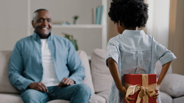Little african american loving daughter hides box gives gift to beloved daddy on birthday or father day congratulates parent surprised happy dad sitting on sofa hugging kid girl strong family embrace - Powered by Adobe