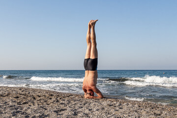 Fototapeta na wymiar Middle age Man in shorts is doing his morning exercises on the beach by the sea. Headstand