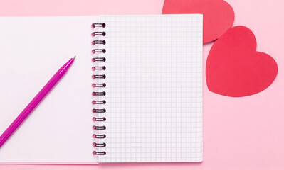 An open blank notepad, a ballpoint pen and two red hearts on a pink background. A love letter. Free space.
