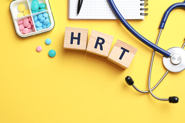 Wooden cubes with abbreviation HRT, pills, stethoscope and notebook on yellow background, flat lay....