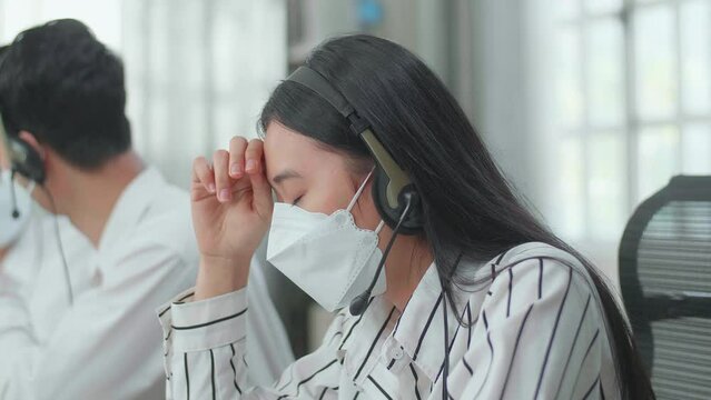 Close Up Of An Asian Woman Call Center Agents Wearing Headset And Mask Headache Because Two Of Her Colleagues Are Talking During Working At The Office
