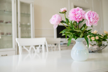 Pink blooming peony flowers with lush green leaves put in vase on white table in light room with modern furniture at home closeup