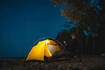 The traveler sits on a camping chair near the glowing tent 