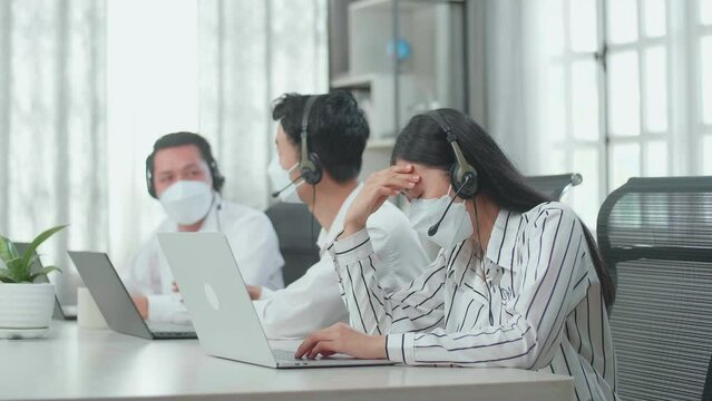 A Woman Of Three Asian Call Center Agents Wearing A Headset, Mask, And Headache Because Two Of Her Colleagues Are Talking With Each Other During Working At The Office

