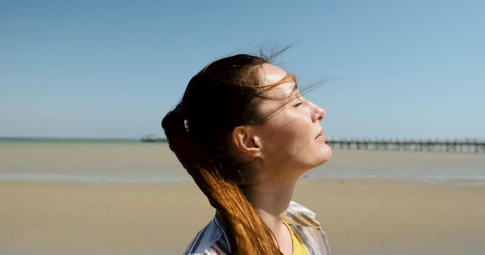 girl with red hair, looks at the bright sun, squinting. Against the background of the sandy tide of the ocean, a clear day, slose up