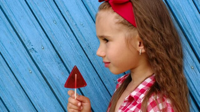 A beautiful long-haired girl with a red bow on head stands sideways in profile and licks a big lollipop. Harm from sweets and the use of large amounts of sugar. Bright juicy combinations