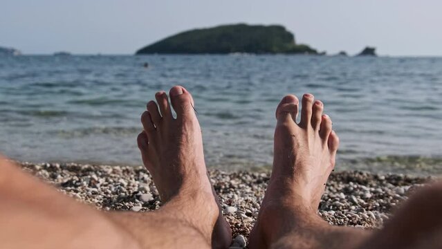 POV view of male barefoot feet on the beach by the sea. Legs of a young man lying on a pebble beach by the calm water. Funny male legs sunbathe and relax on the tropical resort. Summer exotic vacation