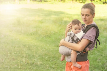 Naklejka na ściany i meble Mom and child on a walk in the park. Mom carries the baby in an ergo bag. Babywearing.