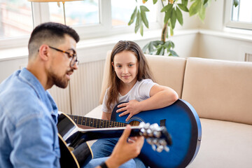 Family time. Dad and daughter together at home. Cute little girl and father playing guitar, sitting on sofa. Caucasian kid child in casual wear learning how to play guitar, perfrom music.