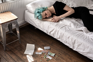 pills spread on floor, young female lying on bed after taking pills overdose, at home. depressed green haired lady in black clothes having hard break up with boyfriend, need mental help