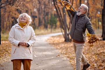 Fototapeta premium Fall season. Family mature couple throwing leaves in autumn forest, having fun outdoors enjoying nature. Elderly Mature Married man and woman in coats spend time together. leisure, lifestyle concept