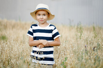 Small farm boy, smiling and looking at the camera, stands in an oat field. Cute boy in a straw hat and a striped T-shirt stands in a field with oats