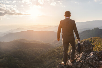 Successful businessman holding his business attache on top rock mountain sunset background, ...