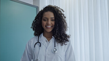 Latin young female doctor wear white uniform, white medical coat, stethoscope and looking at camera