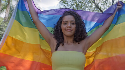 Young curly hair woman covering with lgbt pride flag. Keeping fist up, covering LGBT+ flag