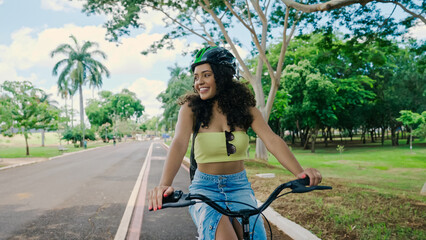 Young Latin woman in protective helmet is riding her bicycle along the bike path in a city park