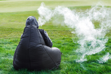 A man smokes a vape, letting off steam from his mouth, sitting on a cushion chair in a field. - obrazy, fototapety, plakaty