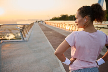 Fototapeta na wymiar Rear view of a pretty athletic woman, female runner athlete, standing with her hands on waist on the city bridge treadmill and looking away, ready for morning cardio training and jogging at sunrise