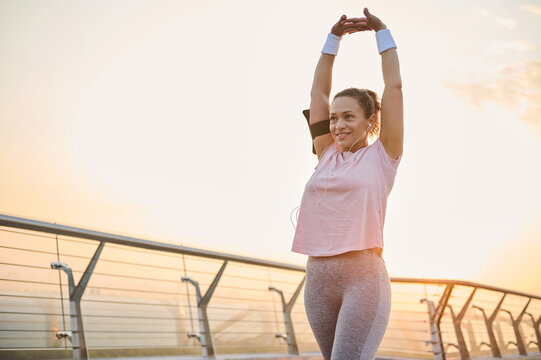 Happy confident female athlete, sportswoman enjoying morning cardio workout, stretching her arms, raising them up, exercising at sunrise. Fitness, health, body care, sport and active lifestyle concept