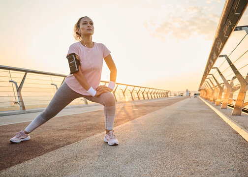 Happy female athlete in activewear enjoying workout in urban environment while doing stretching exercise. Sportswoman stretching legs on a treadmill on a city bridge while working out outdoor