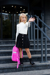 Obraz na płótnie Canvas Blonde girl goes to study. Waving and smiling. In uniform and with a school bag. Primary school, first grader, school student.