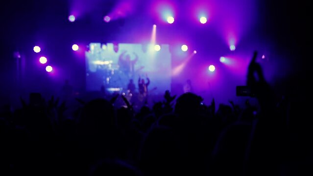 Illuminated stage lights. Raised hands are visible. Concert crowd silhouettes