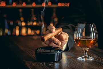 Man's hands with a cigar, elegant glass of brandy on the bar counter. Alcoholic drinks, cognac,...