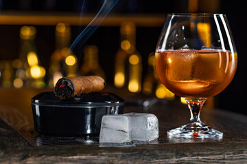 Cigar, elegant glass of brandy on the bar counter. Alcoholic drinks, cognac, whiskey, port, brandy, rum, scotch, bourbon. Vintage wooden table in a pub at night