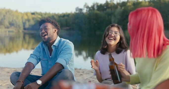 A group of friends is sitting on the beach by the lake, drinking beer by the fire, singing songs, swinging from side to side to the rhythm of music, people of different nationalities