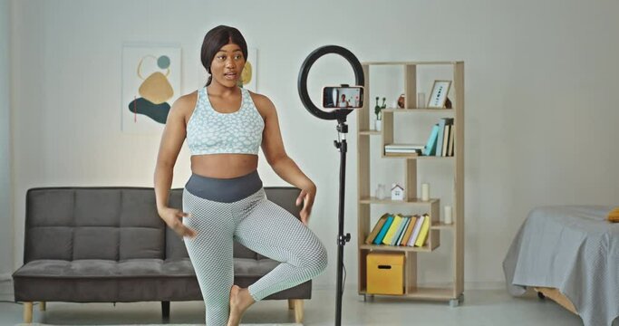 Black curvy woman shooting video and doing yoga. Cheerful plus size African American female yoga instructor recording video on smartphone and standing in Tree pose at home