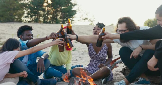 Team of friends is sitting by lake on sand, beach the last days of vacation, they spend time together around the campfire, different nationalities, drink beer in bottles tap them cheers celebrate