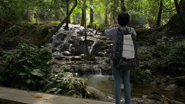 Asian woman tourist with backpack standing on wooden bridge and taking picture with hidden beautiful cascade in tropical rainforest during summer. Travel explorer. Solo camping.