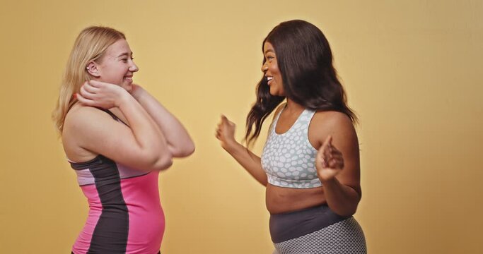 Diverse plump sportswomen complimenting each other. Happy plus size black woman in sportswear smiling and flattering pleased blond girlfriend after fitness training against yellow background