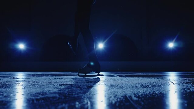 Young female figure skater is performing a woman's single skating choreography on ice rink against soft blue light. Legs artistic skater, practicing sliding and rotation on ice. Slow motion. Close up.