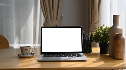 Computer laptop with white blank screen, coffee cup an houseplant on wooden table.