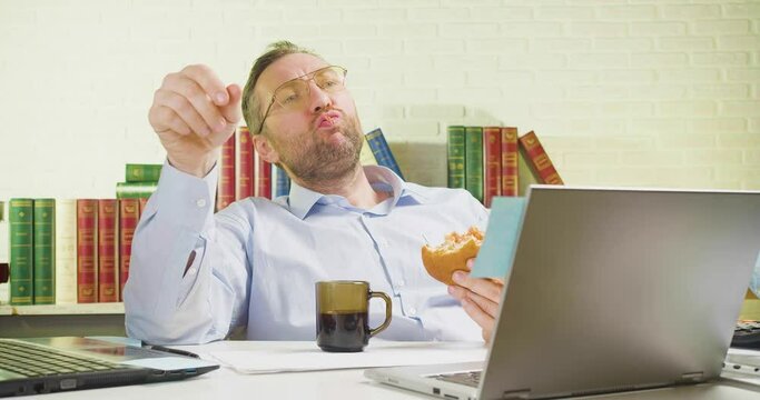 Middle-aged man is lounging in the office - he eats unhealthy food, makes a paper airplane and flies it.