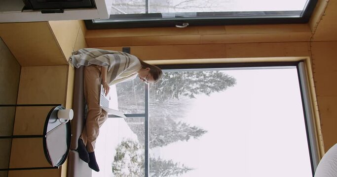 a girl in glasses with collected hair and a plaid on her shoulders sits near the window opens laptop on her knees and types against the backdrop of a winter forest. vertical video. 9:16. 