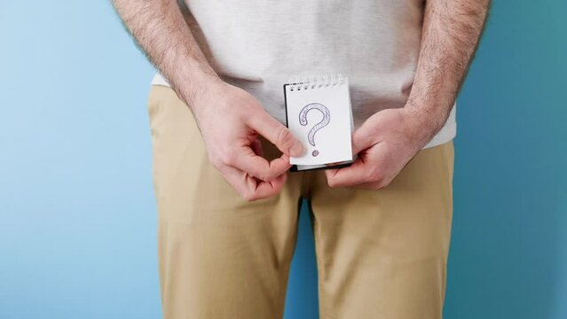 Diseases of the male reproductive system. A man in beige jeans holds a notebook with a question mark in two hands. Close up. Blue background. The concept of artificial insemination and impotence