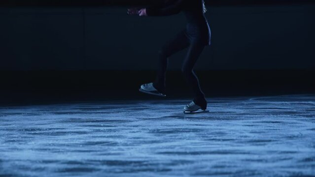 Figure skating lady is skating on ice rink, training at night in the rays of blue light. Young woman preparing to competition, training jumping and slides skills. Slow motion, legs in skates close up.